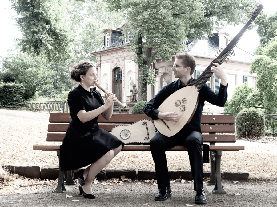 Bild zeigt das Duo Flauto Attiorbato auf einer Bank sitzend in einem Schlosspark. Kathrin Hrtel spielt eine Ganassi-Altblockflte, Konrad Hauser eine Theorbe. Auf der Bank liegt hinter ihnen eine Barockgitarre. Im Hintergrund ist ein Gebude des Bolongaropalastes zu sehen.